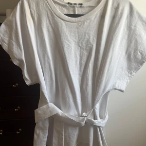 Vit tshirt med bälte från Zara - Säljer en vit tshirt från Zara i mjuk bomull. T-shirten har rund hals, korta ärmar och ett snyggt bälte i midjan som kan knytas för att ge extra form. Perfekt för dig som gillar enkel och clean stil.