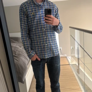 Rutig blå skjorta från Ralph Lauren - Snygg rutig skjorta från Ralph Lauren i blått, vitt, gult och rött. Klassisk button-down med lång ärm, knappar framtill och gul broderad logga på bröstet. Skjortan är i bomull och har en normal passform. Perfekt för dig som gillar preppy stil. Jag väger 75kg och är 185cm💯
