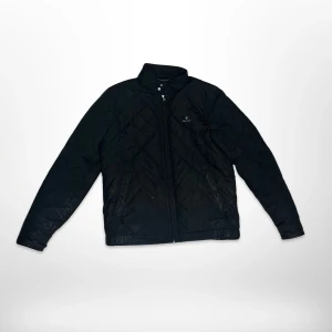 Svart quiltad jacka från Gant - Svart quiltad jacka från Gant med diskret logga på bröstet. Jackan har dragkedja framtill, ståkrage och långa ärmar med knappdetaljer vid ärmslut. Perfekt för dig som gillar stilren och klassisk look.