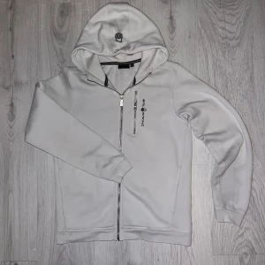 Vit hoodie från Sail Racing med dragkedja - Snygg vit hoodie från Sail Racing med hel dragkedja och broderad logga på bröstet. Tröjan har huva, lång ärm och en extra bröstficka med dragkedja. Perfekt för dig som gillar stilren och sportig look.