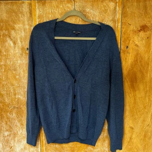 Mörkblå merinoullskofta från Gap - Säljer en stilren mörkblå kofta från Gap i mjuk merinoull. Koftan har v-ringning, knäppning framtill med svarta knappar och långa ärmar. Perfekt att slänga över en t-shirt eller skjorta för en chill look.