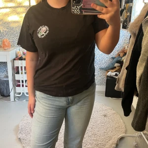 Svart Vans t-shirt med rosprint - Svart t-shirt från Vans med coolt ros-tryck och text både fram och bak. Klassisk rund hals och korta ärmar. Materialet är mjuk bomull och passformen är normal. Perfekt för dig som gillar streetwear och snygga prints.