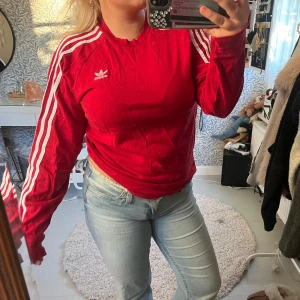 Röd Adidas långärmad tröja - Säljer en röd långärmad Adidas tröja med klassiska vita ränder längs ärmarna och Adidas-logga på bröstet. Tröjan har en rund hals och är gjord i mjuk bomull, perfekt för en sportig och chill stil. Passformen är normal och den är riktigt skön att ha på sig.