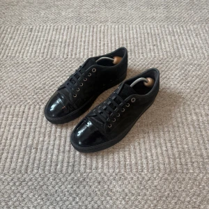 Lanvin Sneakers - Snygga svarta sneakers från Lanvin med ovandel i mocka och lackad tå. Skorna har snörning med metallöljetter och en platt sula i gummi. Perfekta för dig som gillar stilrena och lyxiga detaljer.