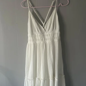 Vit långklänning från Gina Tricot - Säljer en vit klänning från Gina Tricot med smala axelband och v-ringad front. Klänningen har ett plisserat tyg och en lätt volangdetalj nedtill. Perfekt för sommarens alla tillfällen, superfin och luftig design. Storlek L men sitter som en S!!!