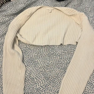 Vit ribbad croppad kofta - Säljer en vit ribbad croppad kofta med lång ärm. Tröjan är stickad och har en tight passform som sitter snyggt över axlarna. Perfekt att slänga över ett linne eller klänning för en chill och trendig look.