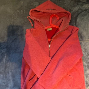 Röd hoodie från Jack & Jones - Säljer en röd hoodie från Jack & Jones med dragkedja och huva. Tröjan har långa ärmar, ribbade muddar och praktiska fickor framtill. Perfekt för chill dagar och enkel att matcha med jeans eller joggers. Snygg och bekväm stil för dig som gillar streetwear.