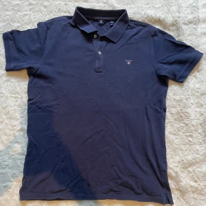 Marinblå GANT pikétröja, 176 cm - Snygg marinblå pikétröja från GANT med klassisk krage och två knappar framtill. T-shirten har korta ärmar och liten GANT-logga broderad på bröstet. Tillverkad i mjuk bomull som är skön mot huden. Perfekt för en chill och stilren look.