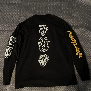 Svart longsleeve från Polar Skate Co - Svart longsleeve tröja från Polar Skate Co i bomull med unika vita grafiska tryck på ryggen och ena ärmen samt gul logga på andra ärmen. Perfekt för dig som gillar streetwear och vill sticka ut med coola detaljer. Tröjan har varit använd väldigt fåtal gånger och har inga defekter
