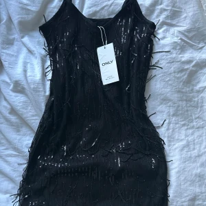 Svart paljettklänning från ONLY XS - Supersnygg svart kort klänning från ONLY med tunna axelband och massor av glittriga paljetter. Klänningen har en figurnära passform och coola detaljer med hängande paljetttrådar som ger extra partykänsla. Perfekt för dig som vill sticka ut!