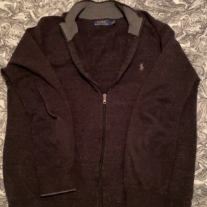 Tröja Ralph Lauren  - Mörkgrå/svart kofta från Polo Ralph Lauren med hel dragkedja och diskret logga på bröstet. Tröjan har ribbade muddar, hög krage i kontrastfärg och är långärmad. Perfekt för lager-på-lager och chill stil.