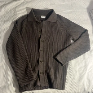 Brun ribbad kofta från John Henric - Säljer en brun ribbstickad kofta från John Henric i storlek S. Koftan har klassisk krage, knäppning framtill med fem knappar och långa ärmar. Perfekt för lager-på-lager och chillare dagar.