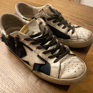 Golden Goose sneakers - Snygga sneakers från Golden Goose med vit skinnbas, svarta snören och mörkblå detaljer i tyg. Ikonisk vit stjärna på sidan och slitna kanter för en cool, vintage look. Perforerad tå och platt sula ger en klassisk streetstyle-vibe. Slitningen på skorna är själva designen. 
