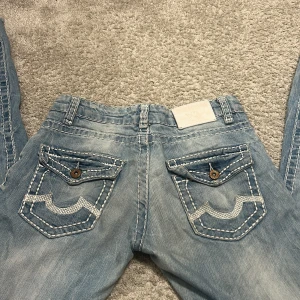 True Religion ljusblå jeans, stl 27 - Säljer ett par ljusblå jeans från True Religion med snygga slitningar och kontrastsömmar. Jeansen har klassiska bakfickor med broderad detalj och knappar. Modellen är rak med låg midja och tillverkad i mjukt bomullstyg. Aldrig använda och ser kanske inte så bootcut, men det är för att de är för små för mig. Storlek 27 ❤️