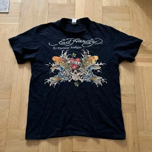 Svart Ed Hardy t-shirt med tryck - Svart t-shirt från Ed Hardy med stort färgstarkt tryck på bröstet som visar ett hjärta, dödskalle och fiskar. Texten 'Love Kills Slowly' och Ed Hardy-loggan syns tydligt. Klassisk rund hals och korta ärmar. Materialet är mjuk bomull. Har några defekter därav pris (kom dm för bilder)