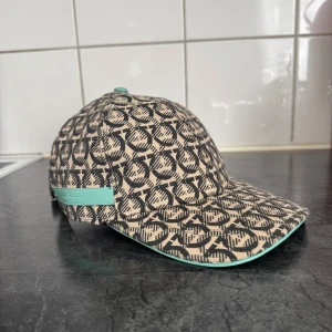 Beige och svart mönstrad keps Salvatore Ferragamo - Snygg keps från Salvatore Ferragamo med beige och svart grafiskt mönster över hela kepsen. Justerbar rem med metallspänne och en mintgrön detalj baktill samt under skärmen. Perfekt för dig som vill sticka ut med en unik accessoar.