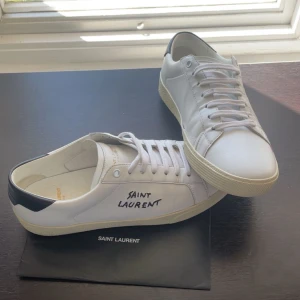 Saint Laurent vita sneakers i skinn - Snygga vita sneakers från Saint Laurent med svart hälparti och handskriven logga på sidan. Säljer dom för att har tröttnat lite på dom och är sugen på något nytt! 8/10 i skick, sulan på höger skon är lite sliten. Ny pris: 5000-6000kr mitt pris:2099kr. Hör av er om ni har några frågor!////Ville