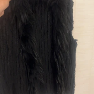 Svart pälsväst med stickad bas - Säljer en svart pälsväst med stickad bas och långa, mjuka pälsdetaljer framtill. Västen har en snygg struktur med blandning av stickat och päls. Perfekt att slänga över en hoodie eller långärmad tröja för extra edge.