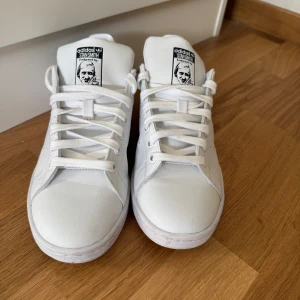 Adidas Stan Smith vita sneakers - Klassiska Adidas Stan Smith sneakers i vitt skinn med svarta detaljer på hälen. Snygg minimalistisk design med perforerade ränder på sidorna och vit platt sula. Skorna har snörning och rund tå, perfekt för en clean och stilren look.