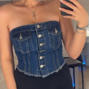 Jeans bandeau tubtopp med knappar - Säljer en snygg jeans bandeau tubtopp med silverfärgade knappar framtill. Toppen är croppad och har smock baktill för extra passform. 