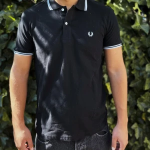 Svart Fred Perry piké med vit kant - Svart pikétröja från Fred Perry med klassisk vit broderad logga på bröstet. Tröjan har vit kant på krage och ärmslut, två knappar framtill och är gjord i mjuk bomull. Perfekt för dig som gillar stilren och sportig look.  .     Nypris: 1099kr GORILLAPRIZE:739kr             Skick:10/10 oöppnad          KVITTO MEDKOMMER   