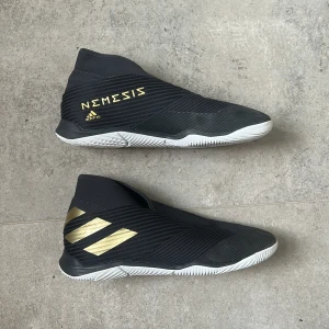 Adidas Nemeziz svarta fotbollsskor - Svarta Adidas Nemeziz fotbollsskor med guldiga detaljer och vita sulor. Skorna har en strumpliknande passform utan snörning och är tillverkade i syntetmaterial. Perfekta för dig som vill ha stilrena och moderna skor på planen.