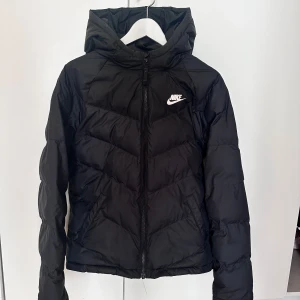 Svart pufferjacka från Nike med huva - Snygg svart pufferjacka från Nike med huva och dragkedja framtill. Jackan har ett stilrent Nike-logo på bröstet och quiltad design som ger en cool look. Perfekt för kalla dagar när du vill hålla stilen och värmen.