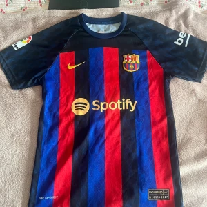 FC Barcelona Lewandowski matchtröja - Säljer en officiell FC Barcelona matchtröja från Nike med Lewandowski och nummer 9 på ryggen. Tröjan har klassiska röda och blå vertikala ränder, guldiga detaljer och sponsorloggor som Spotify och UNHCR. Kortärmad modell i lätt och ventilerande material. Köpt i Spanien 🇪🇸 