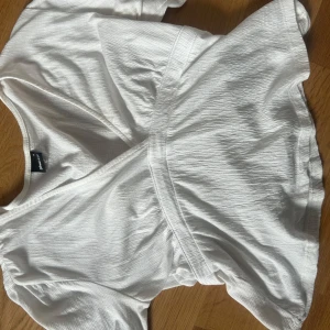 Vit omlottopp från Gina Tricot - Säljer en vit omlottopp från Gina Tricot i storlek S. Toppen har långa ärmar, knyt i ryggen och en v-ringad design. Materialet är lätt strukturerat och känns luftigt, perfekt för en fräsch look. Passar dig som gillar stilrena och enkla plagg.Säljs ej längre.