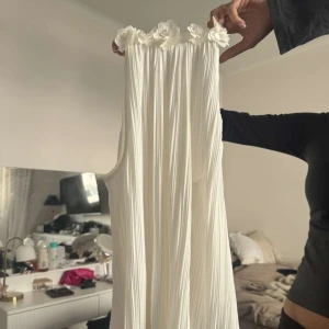 Vit legalt  kort topp med blommor - Supersöt vit elegant topp  med plisserat tyg och dekorativa blommor längs halsringningen. Toppen är ärmlös och har en luftig, flowy passform som känns både trendig och romantisk. Perfekt för sommarens alla tillfällen! Aldrig andvänd, väldigt fin