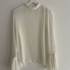 Vit plisserad blus med volangärm - Superfin vit blus i plisserat tyg med högt hals med volangkant och långa ärmar som avslutas med bred volang och knappdetalj. Blusen har en lätt transparent känsla och knäppning i nacken. Perfekt för dig som gillar romantiska och luftiga plagg.