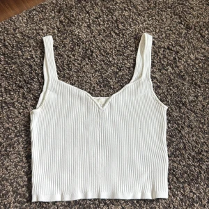 Krämvit stickad croppad linnetopp - Säljer en croppad linnetopp med breda axelband och v-ringning framtill. Toppen är ribbad och har en enkel, stilren look som passar perfekt till jeans eller kjol. Materialet är mjukt och stretchigt för en bekväm passform.