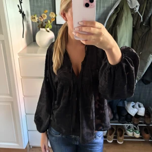 Svart blus med voluminösa ärmar - Säljer en svart blus med voluminösa, långa ärmar och v-ringad hals. Blusen har ett subtilt mönster i tyget och en luftig, loose passform som ger en cool och avslappnad vibe. Perfekt att styla med jeans eller kjol för en trendig look.