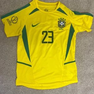 Brasilien Kaka #23 Nike fotbollströja - Gul Brasilien fotbollströja från Nike med gröna detaljer och nummer 23, samt Kaka' på ryggen. Broderat CBF-emblem och Nike-logga på bröstet. Kortärmad modell i lätt och ventilerande polyester, perfekt för match eller träning. Storlek S passar kanske M