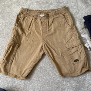 Beige cargoshorts från H&M - Säljer ett par beige cargoshorts från H&M med elastisk midja och snörning. Shortsen har stora sidofickor med lock och en cool New York City-detalj på benet. Tillverkade i mjuk bomull, perfekta för chill dagar och streetstyle.