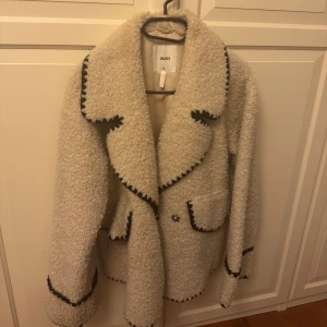 Beige teddyjacka från Object - Säljer en beige teddyjacka från Object med svarta dekorativa sömmar längs kanterna. Jackan har stora slag, två fickor framtill och en boxig, lite oversized passform. Perfekt för att hålla sig varm och snygg under höst och vinter! Obs: bilden från google är en annan lite mörkare färg då färgen jag köpt inte längre verkar finnas🤍 ord pris: 1199 kr