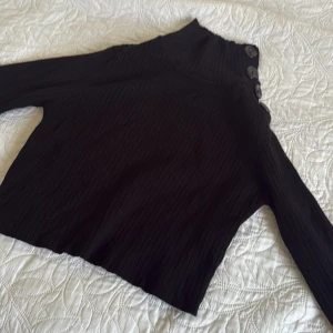 Svart croppad och ribbad tröja med knappar - Svart polotröja, croppad topp med ribbad struktur och stora knappar längs halsen. Tröjan har lång ärm och är gjord i ett stickat material som känns mjukt och skönt. Den kan användas som XS/S