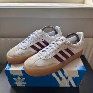 Adidas Samba vit/bordeaux 42 2/3 - Adidas Samba sneakers i vitt skinn med bordeauxfärgade ränder och beige mockadetaljer. Klassisk gummisula och snörning framtill. Perfekt för dig som gillar retrostil och vill ha en ikonisk sneaker med snygga kontraster.