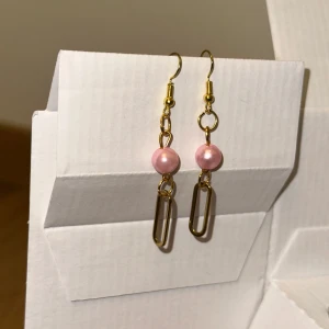 Guldiga örhängen med rosa pärlor - Snygga örhängen med guldiga detaljer och en rund rosa pärla i mitten. Hängande design med en avlång länk under pärlan som ger ett coolt och modernt uttryck. Perfekta för att lyfta din stil med en touch av färg och elegans.