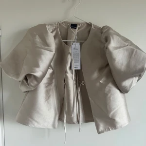 Champagnefärgad puffärm blus Gina Tricot - Supersnygg blus i champagnefärg från Gina Tricot med glansigt satinliknande material. Blusen har stora puffärmar, knytband framtill oChampagnefärgad puffärm blus Gina Tricotch en rak, lite croppad passform. Perfekt för dig som vill sticka ut med en trendig och elegant look.