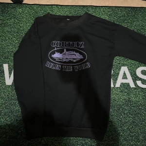 Svart Corteiz sweatshirt med tryck - Svart sweatshirt från Corteiz med lila tryck på bröstet där det står 'Corteiz Rules The World' och en grafisk illustration. Tröjan har rund hals och långa ärmar.
