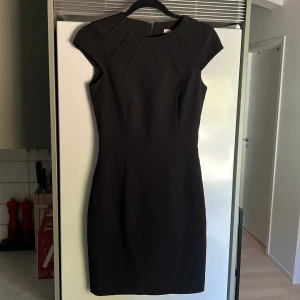 Svart kortklänning med struktur - Snygg svart kortklänning med coola strukturerade detaljer vid axlarna och en rund halsringning. Klänningen har en figurnära passform och korta ärmar, perfekt för dig som gillar stilrena och eleganta plagg. Dragkedja bak för enkel på- och avtagning.