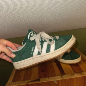 Adidas Campus gröna sneakers mocka - Gröna Adidas Campus sneakers i mocka med klassiska vita ränder på sidorna och vit sula. Skorna har rund tå, platt sula och snörning. Perfekta för dig som gillar retrostil och streetwear.