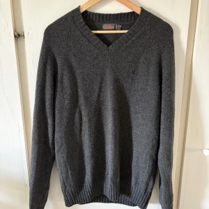 Mörkgrå stickad tröja från Morris M - Mörkgrå stickad tröja från Morris med v-ringad hals och diskret broderad logga på bröstet. Tröjan har långa ärmar och ribbade muddar vid hals, ärmslut och nederkant. Perfekt för dig som gillar klassisk stil med en modern touch.