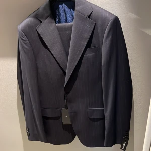 Mörkblå randig kavaj från Sand - Snygg mörkblå kavaj från Sand Copenhagen med diskreta vita ränder. Klassisk passform, fyra knappar vid ärmslut och två fickor med lock framtill. Tillverkad i 100% ny ull för en lyxig känsla. Perfekt för dig som vill ha en stilren och modern look.