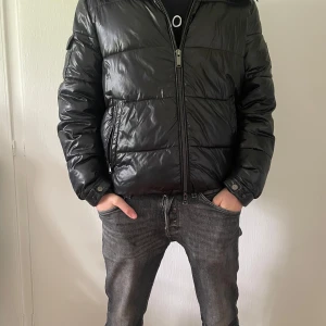 Svart pufferjacka från Michael Kors - Svart pufferjacka från Michael Kors med glansig finish och huva. Jackan har quiltad design, dragkedja framtill och sidofickor med tryckknapp. Märkespatch på ärmen ger extra stilpoäng. Perfekt för kalla dagar när du vill hålla dig varm