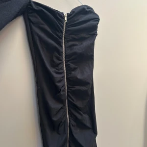 Svart axelbandslös klänning med dragkedja - Snygg svart axelbandslös kortklänning med dragkedja framtill och rynkade detaljer längs hela klänningen. Perfekt för dig som vill sticka ut med en edgy look. Figurnära passform och cool design.