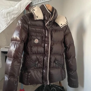 Moncler jacka - Säljer en mörkbrun jacka från Moncler med vita detaljer på kragen och insidan. Jackan har dragkedja, fickor med dragkedja och knapp, samt Moncler-logga på bröstet. Perfekt för kalla dagar och riktigt snygg till jeans. Riktigt bra skick.