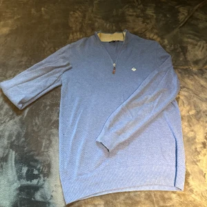 Blå tröja med half zip från Sir Raymond Tailor - Snygg blå långärmad tröja från Sir Raymond Tailor i storlek M. Tröjan har ett diskret mönster, ribbade muddar och krage i kontrastfärg. Half zip framtill med läderdetalj på dragkedjan och broderad logga på bröstet. Perfekt för chill dagar.