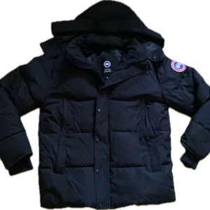 Svart pufferjacka från Canada Goose - Svart pufferjacka från Canada Goose med ikonisk patch på ärmen. Jackan har huva, dragkedja och knappar framtill samt två sidofickor. Fylld med dun och tillverkad i slitstark polyester, perfekt för kalla vinterdagar. Snygg och klassisk look med loose passform.
